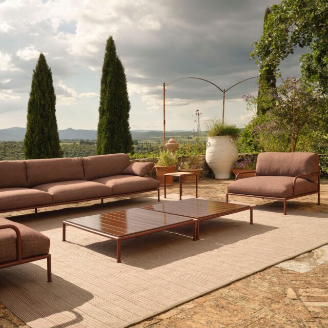 Außenterrasse mit braunen gepolsterten Sitzmöbeln, einem Couchtisch aus Holz und einem großen Teppich auf Steinboden. Umgeben von hohen grünen Bäumen, zeigt dieses Gartendesign fachmännischen Garten- und Landschaftsbau vor einer malerischen Kulisse aus Hügeln und bewölktem Himmel.