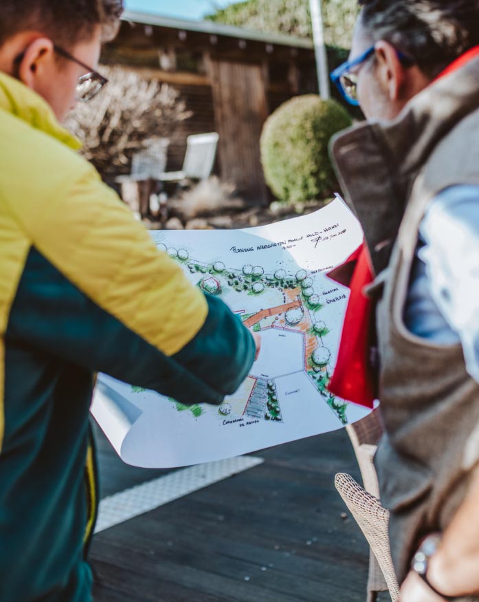 Zwei Personen betrachten im Freien einen detaillierten Garten- und Landschaftsbauplan, auf dem Bäume, Wege und beschriftete Abschnitte eingezeichnet sind. Eine Person zeigt auf den Plan, und beide tragen Jacken, die von der in Luxemburg und Deutschland geschätzten Kompetenz zeugen.