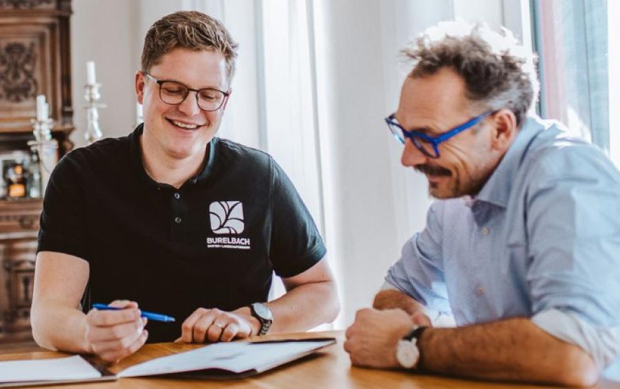 Zwei Männer sitzen an einem Tisch und sehen gemeinsam Garten- und Landschaftsbauunterlagen durch. Ein Mann lächelt und hält einen Stift in der Hand, während der andere die Papiere betrachtet. Sie scheinen ein freundliches Gespräch in einem gut beleuchteten Raum zu führen.