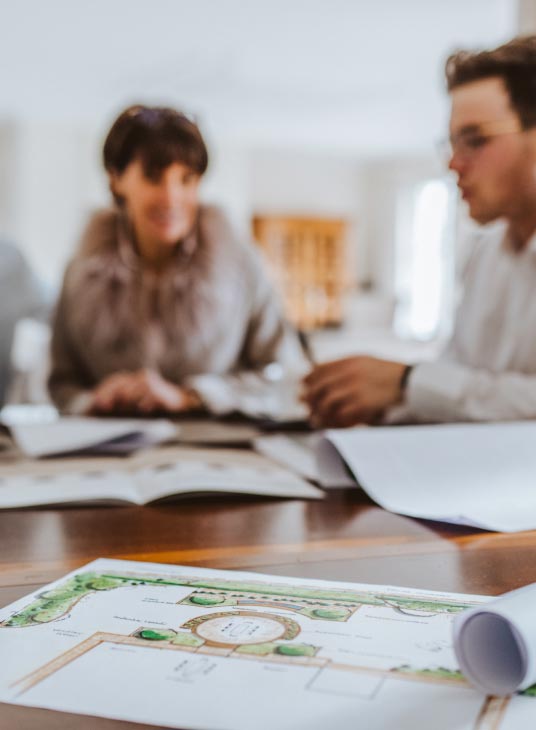 Nahaufnahme einer Grundrisszeichnung auf einem Tisch mit zwei unscharfen Personen im Hintergrund, die in einem gut beleuchteten Burelbach-Büro Dokumente über Gartenmöbel in Luxemburg und Deutschland besprechen.
