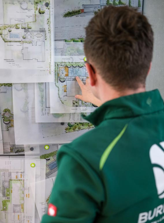 Eine Person in grüner Jacke zeigt auf Architektur- oder Landschaftspläne, die an einer Tafel befestigt sind, und studiert die Entwürfe für Gartendesign und Gartenmöbel genau.
