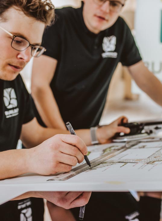 Zwei Personen mit Brillen und schwarzen Hemden arbeiten gemeinsam an architektonischen Zeichnungen. Mit Bleistift und Lineal entwerfen sie handwerklich inspirierte Gartenmöbel auf einem großen Blatt Papier, das auf einem Schreibtisch liegt.
