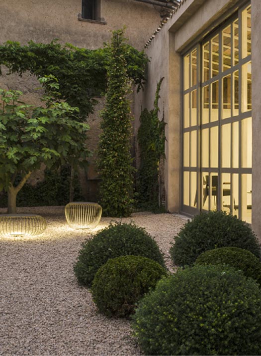 Ein von Burelbach Gardendesign gestalteter Kieshof mit gepflegten grünen Sträuchern, einer hohen efeubewachsenen Säule, einem belaubten Baum und leuchtenden modernen Außenleuchten neben einem großen Gebäude mit Fenstern in Luxemburg und Deutschland.