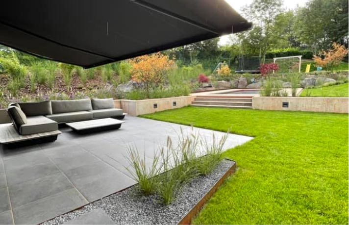 Ein moderner Garten mit einer Terrasse mit grauem Sofa und Couchtisch, umgeben von grünem Rasen, Pflanzen, Steinstufen und einem Fußballtor in der Ferne - ein Musterbeispiel für Gardendesign und Qualitätshandwerk.