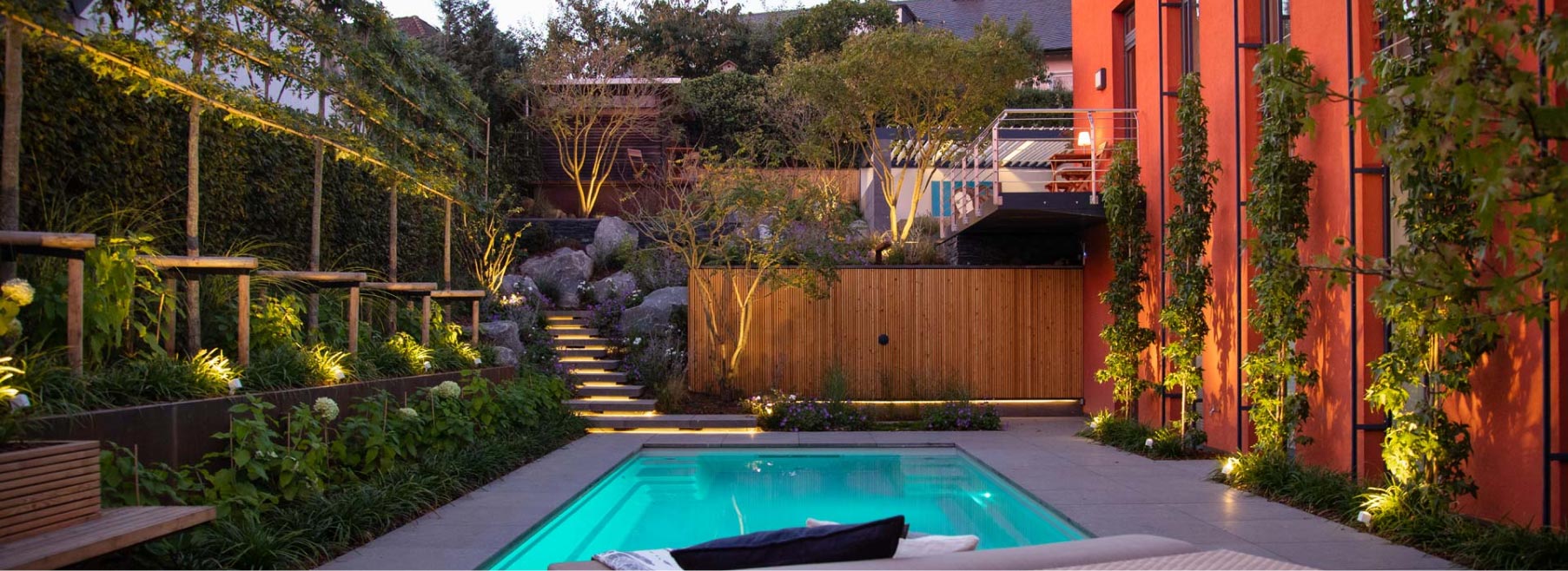 Ein schmaler moderner Hinterhof in der Abenddämmerung mit einem kleinen rechteckigen Pool, üppigen Grünpflanzen entlang einer mit Weinreben bewachsenen Mauer, warmer Beleuchtung, handwerklich inspirierten Gartenmöbeln, einem Holzzaun und Sitzgelegenheiten in der Nähe eines orangefarbenen Gebäudes.