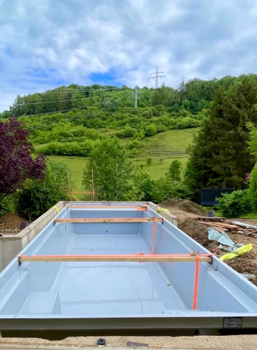 Ein rechteckiger, in den Boden eingelassener Swimmingpool von Burelbach wird gerade in einem Hinterhof gebaut, umgeben von Erde und Grün, mit Hügeln und Bäumen im Hintergrund unter einem teilweise bewölkten Himmel - ein Beispiel für fachmännischen Garten- und Landschaftsbau.