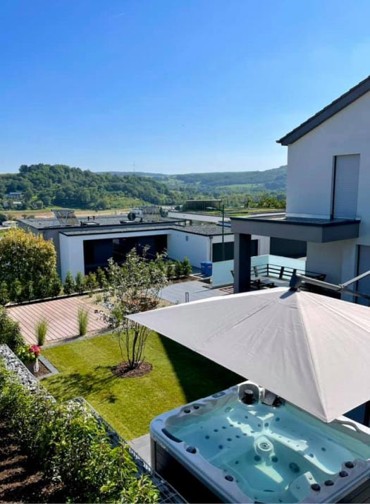 Modernes Haus mit einem Whirlpool unter einem großen Sonnenschirm im Garten, mit Burelbach Garten- und Landschaftsbau auf der Holzterrasse und dem begrünten Hof. Moderne Häuser und die grünen Hügel von Luxemburg und Deutschland vervollständigen die malerische Kulisse.