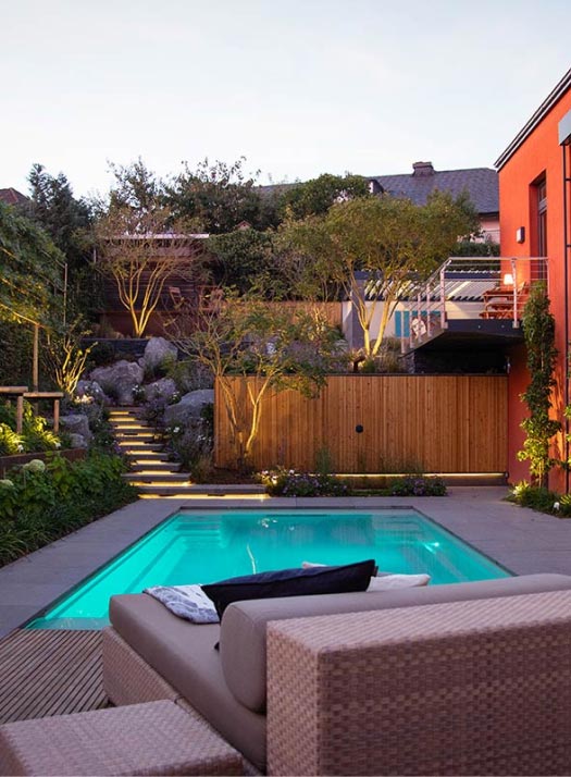 Ein moderner Garten mit einem kleinen rechteckigen Pool, Korbsesseln und üppigen Pflanzen zeugt von fachmännischem Gartendesign. Warme Lichter beleuchten die Steinstufen zu einer erhöhten Terrasse, während eine rote Wand und ein Holzzaun für Privatsphäre sorgen - perfekt für Luxemburg und Deutschland.