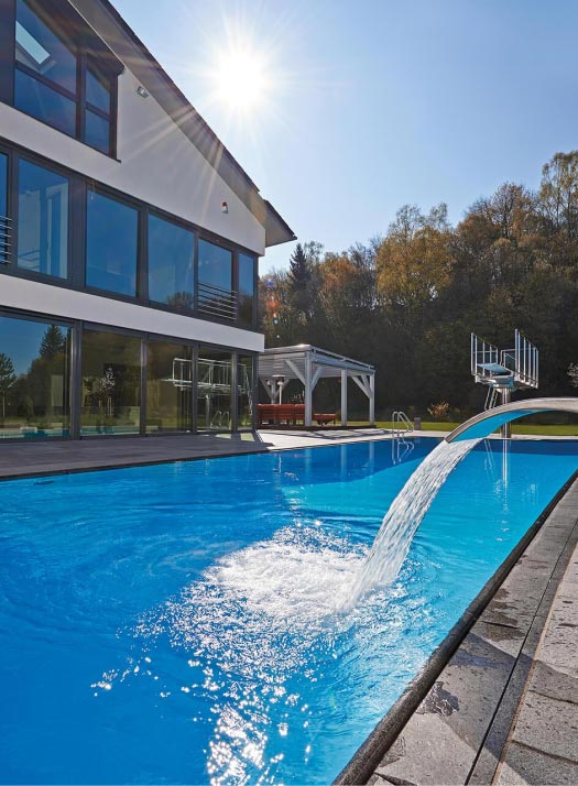 Ein modernes Haus mit großen Fenstern steht neben einem klaren, blauen Swimmingpool, aus dem Wasser in Kaskaden aus einem Springbrunnen fließt - ein Beispiel für exquisites Garten- und Landschaftsbauhandwerk aus Luxemburg und Deutschland. Bäume und ein Pavillon vervollständigen die Szene.