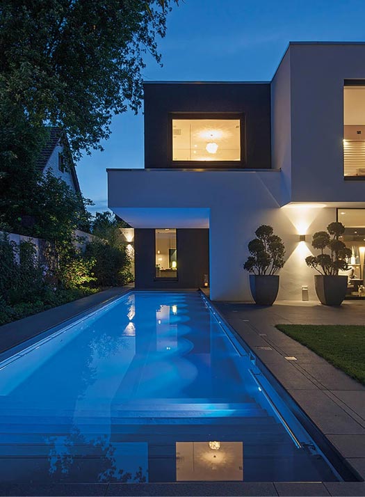 Modernes Haus in der Abenddämmerung mit großen Fenstern und beleuchteten Innenräumen, mit einem langen, schmalen Swimmingpool mit blauen Lichtern und einem von Experten für Garten- und Landschaftsbau gestalteten Garten, perfekt für Gardendesign in Luxemburg und Deutschland.