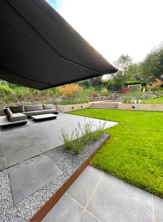Ein moderner Garten von Burelbach Garten- und Landschaftsbau mit einer gefliesten Terrasse, stilvollen Gartenmöbeln mit niedrigen Sitzpolstern, grünem Rasen, terrassenförmiger Bepflanzung mit Stufen, üppigen Pflanzen und Bäumen sowie einer großen Markise als Schattenspender.
