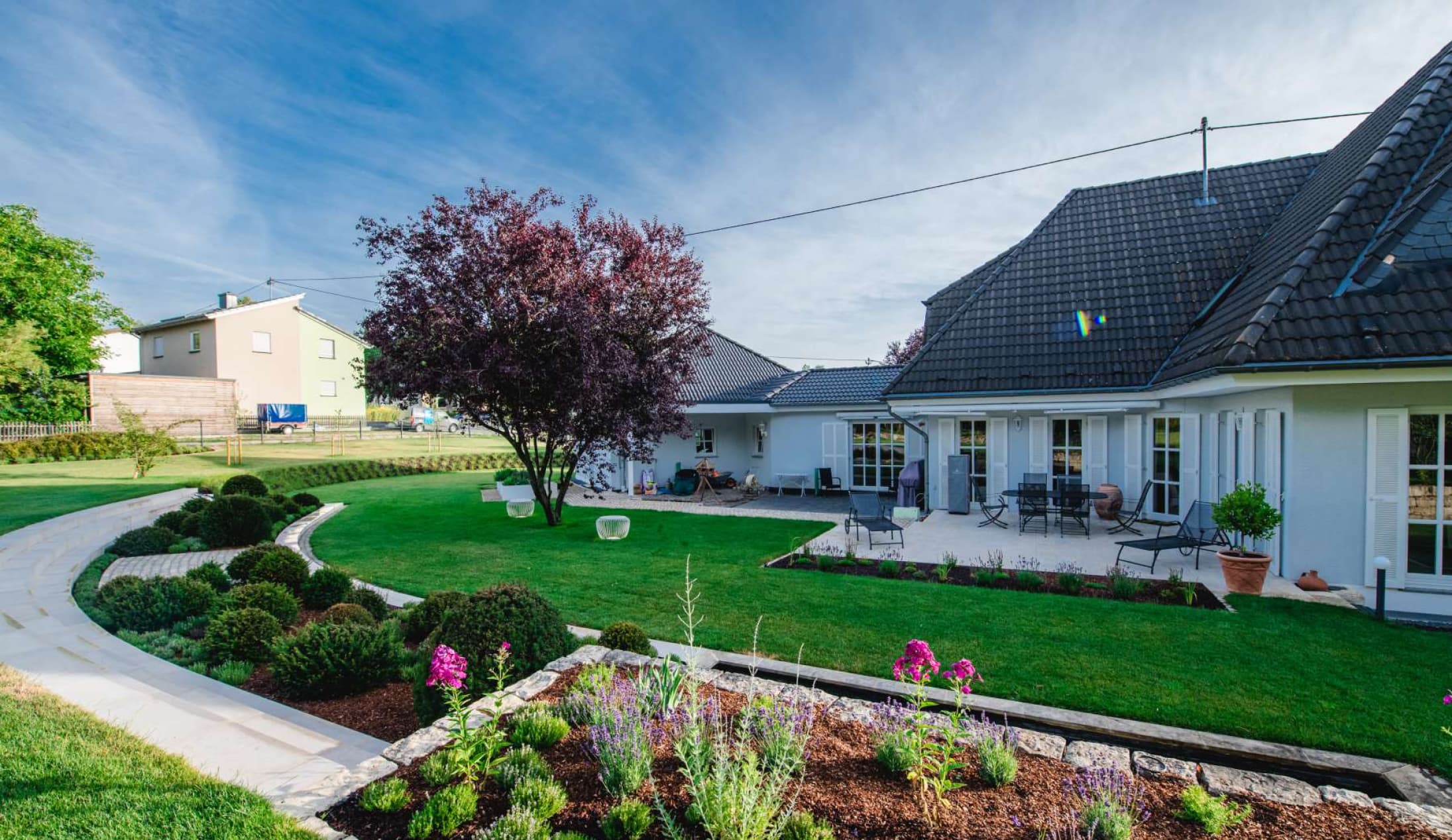 Ein modernes Haus mit grauem Dach und weißen Wänden, mit einer großzügigen Terrasse mit stilvollen Gartenmöbeln, gepflegtem Rasen, geschwungenem Weg, Blumenbeeten und ordentlich gestutzten Sträuchern unter blauem Himmel - perfektes Gardendesign, gesehen in Luxemburg und Deutschland.