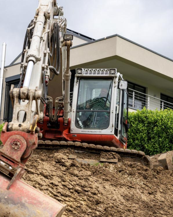 Ein rot-weißer Bagger gräbt Erde vor einem modernen Haus mit großen Fenstern und einem Balkon, umgeben von Grün - eine beeindruckende Szene, die Handwerk in Luxemburg und Deutschland unter einem bewölkten Himmel zeigt.