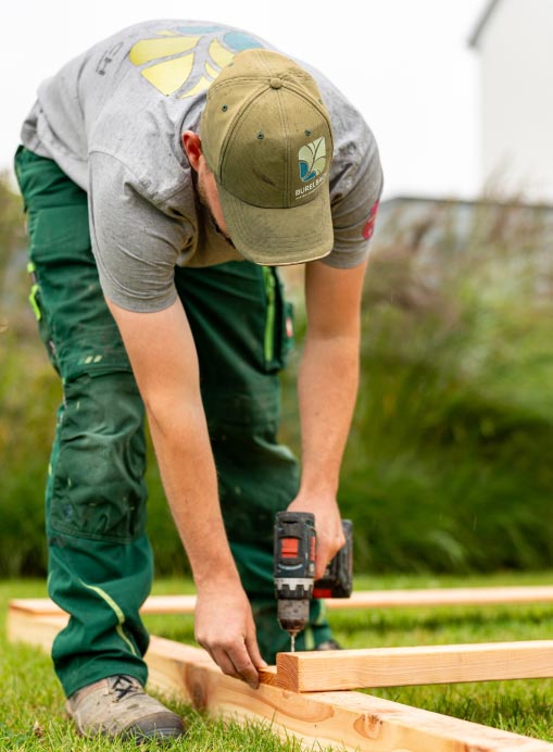 Eine Person mit Mütze, grauem Hemd und grüner Hose setzt mit einer Bohrmaschine Gartenmöbel im Freien zusammen und zeigt auf der Wiese ihr handwerkliches Können.