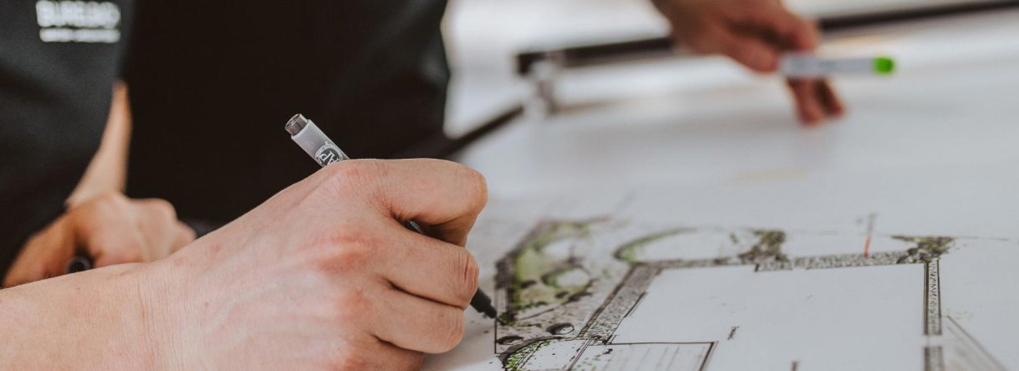 Nahaufnahme der Hand einer Person, die mit einem Stift Architektur- oder Landschaftspläne auf Papier zeichnet - eine perfekte Inspiration für Gartenmöbel-Design und Handwerk in Luxemburg und Deutschland -, während eine andere Hand einen Marker über dem Tisch hält.