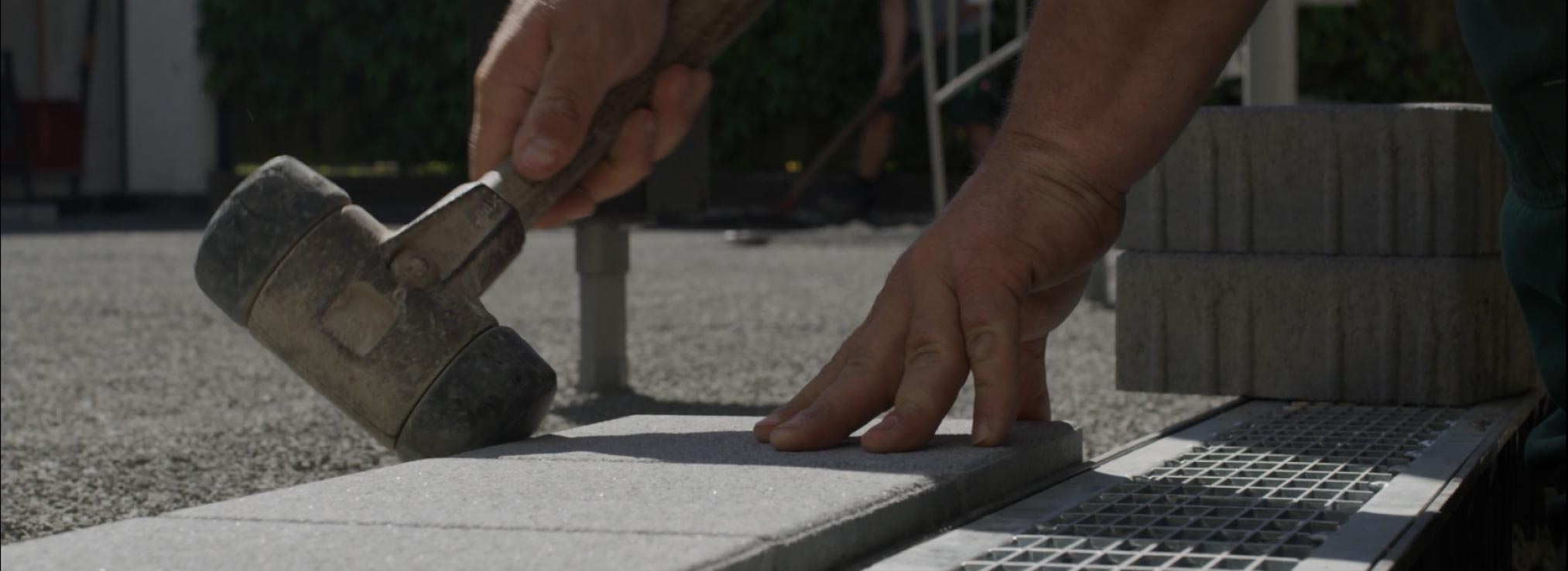 Ein Handwerker klopft mit einem Gummihammer rechteckige Betonpflastersteine auf eine Kiesfläche neben einem Drainagegitter. Gestapelte Pflastersteine und verschwommenes Grün deuten auf den professionellen Garten- und Landschaftsbau von Burelbach hin.