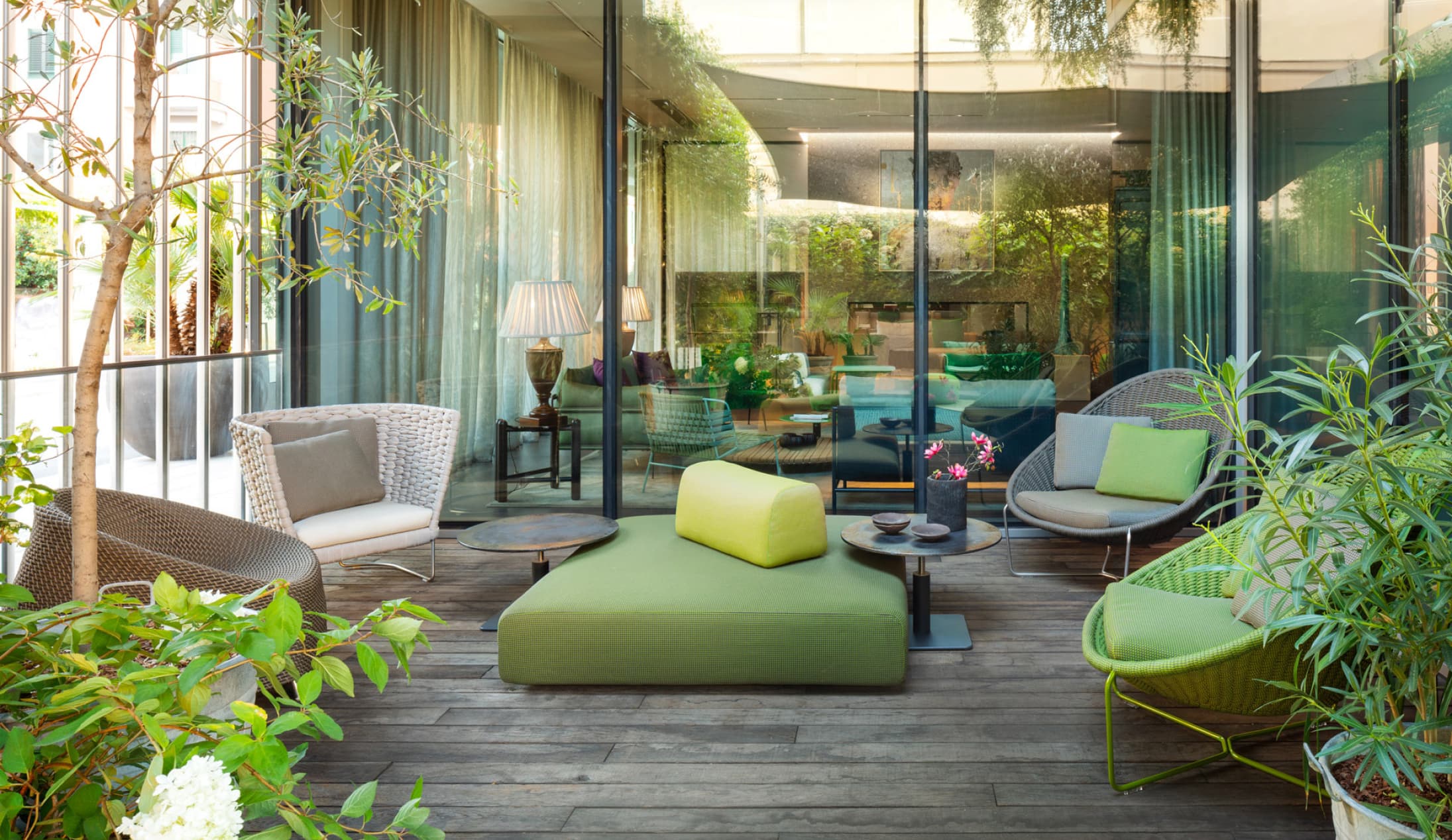 Eine moderne Außenterrasse mit grünen Sitzpolstern, Korbstühlen, kleinen Tischen und üppigen Pflanzen - ein Beispiel für elegantes Gardendesign - steht auf einem Holzboden vor großen Glastüren in stilvollen Häusern in Luxemburg und Deutschland.