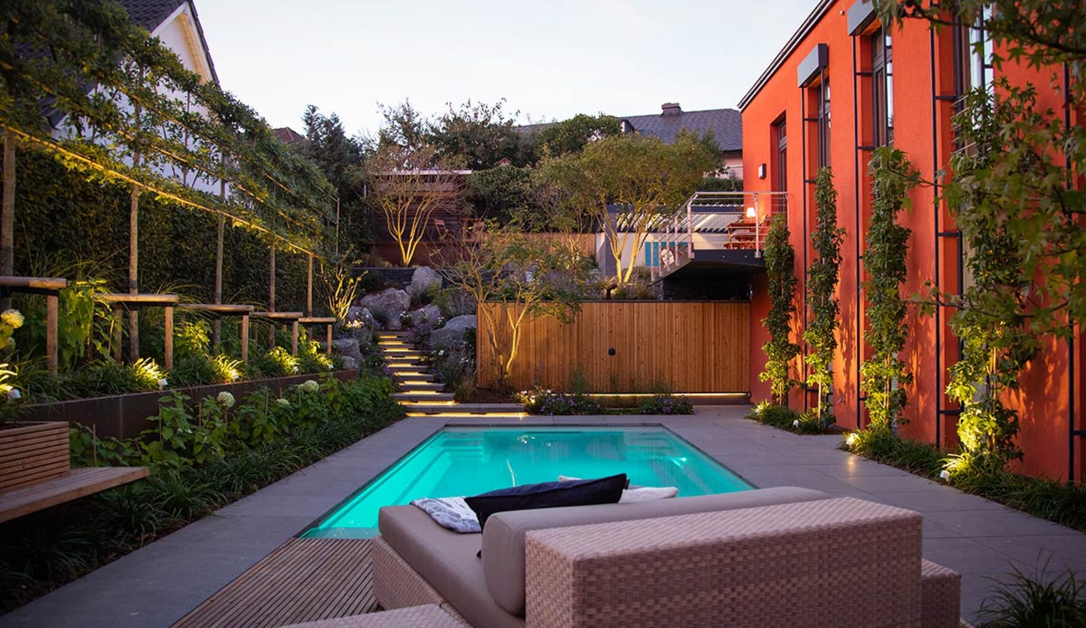 Ein moderner Garten mit rechteckigem Pool von Garten- und Landschaftsbau Profis aus Luxemburg und Deutschland mit üppigem Grün, einem beigen Lounge-Sessel und einer warmen Wegbeleuchtung, die in der Abenddämmerung eine gemütliche, einladende Atmosphäre schafft.