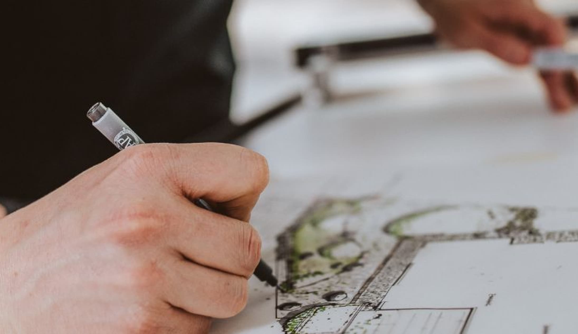 Die Hand einer Person zeichnet mit einem Stift Architektur- oder Landschaftspläne auf ein weißes Blatt Papier, wobei das handwerkliche Geschick hervorgehoben wird, während im Hintergrund ein Teil einer anderen Hand und Zeichenwerkzeuge zu sehen sind.