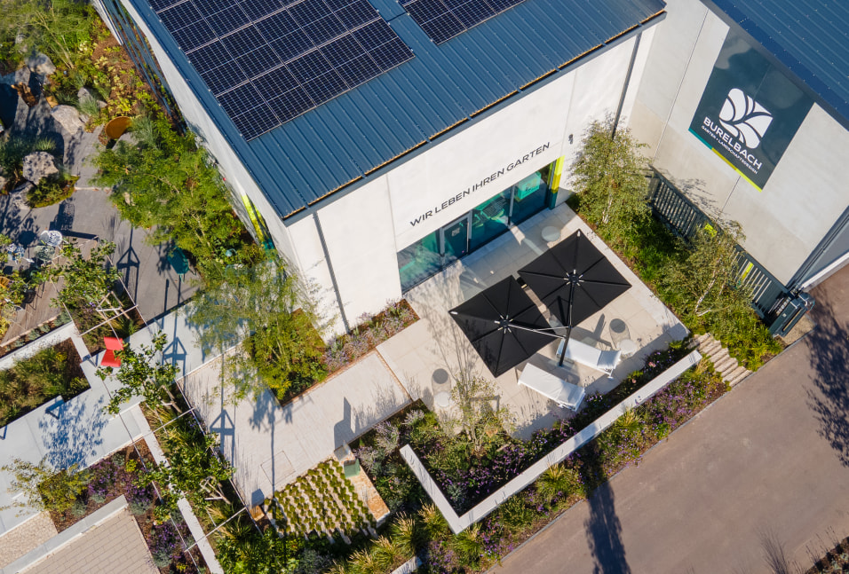Luftaufnahme eines modernen Burelbach-Gebäudes mit Sonnenkollektoren auf dem Dach, einem begrünten Innenhof mit Tischen, Stühlen und Sonnenschirmen sowie üppiger Gardendesign-Begrünung. Ein großes Logo ist an der Außenwand angebracht.