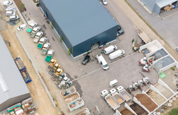 Luftaufnahme einer Industrieanlage mit Lastwagen, Transportern und Baumaterialien sowie Gartenmöbeln, die in offenen Behältern rund um ein großes Lager für Luxemburg und Deutschland gelagert werden, was die dynamische Handwerksindustrie der Region verdeutlicht.