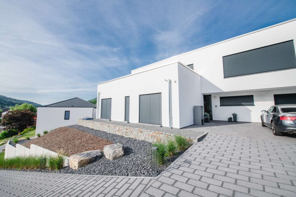 Ein modernes weißes Haus mit großen Fenstern und Metallläden, einer abschüssigen Auffahrt, einer Steinlandschaft, stilvollen Gartenmöbeln auf der Terrasse und einem silbernen Auto, das unter einem blauen Himmel geparkt ist.