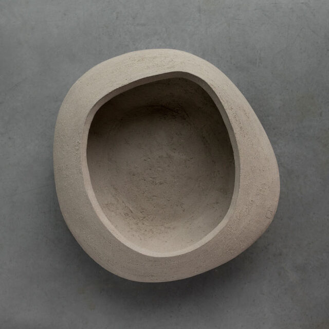 Eine große, asymmetrische, abgerundete Schale aus Stein oder Ton, die mit Handwerk hergestellt wurde, sitzt auf einer glatten grauen Oberfläche. Die Schale hat eine organische, unebene Form und ein natürliches, unfertiges Aussehen.