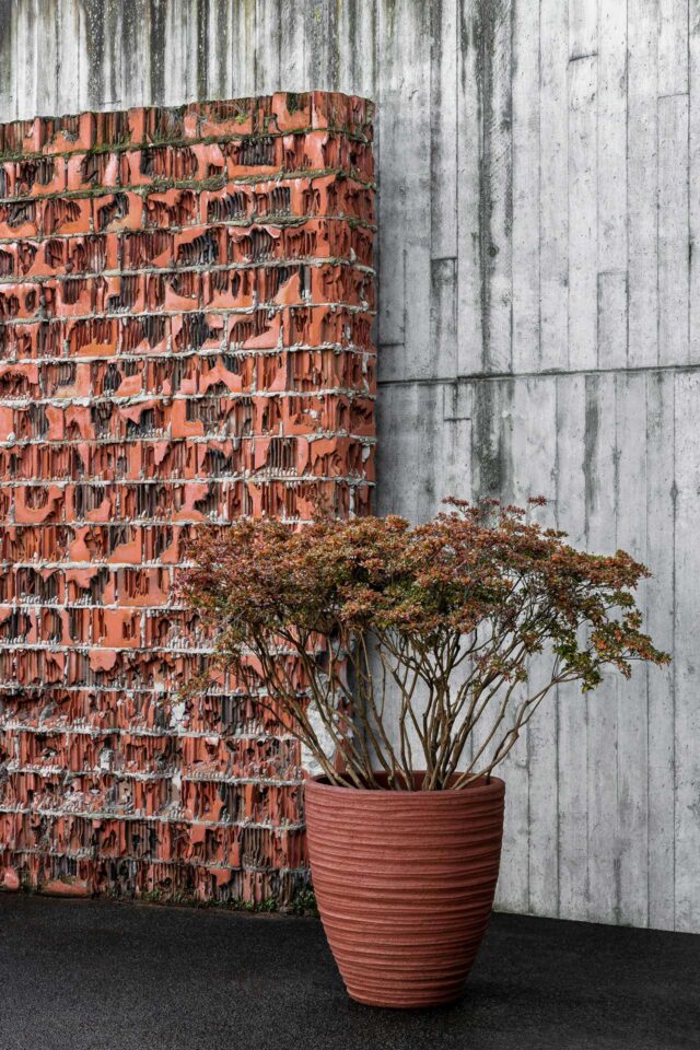 Eine strukturierte rote Ziegeltafel steht vor einer grauen Betonwand, mit einem grünen Strauch in einem großen, geriffelten Terrakottatopf, der Handarbeit und durchdachte Gartengestaltung zeigt, auf dunklem Grund im Vordergrund.