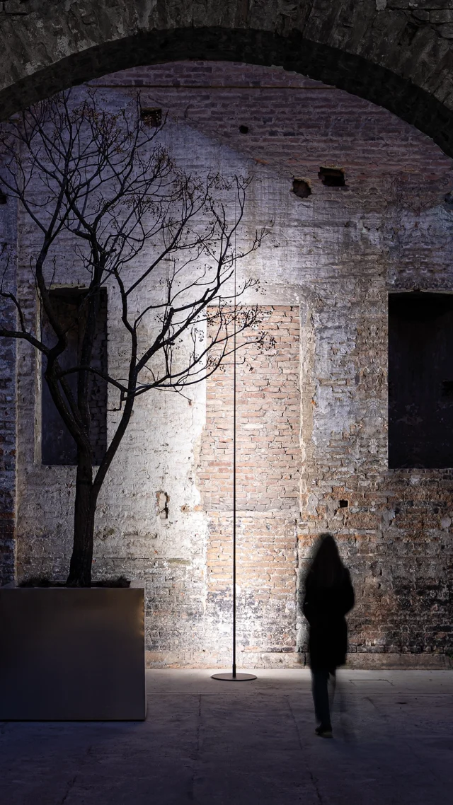 Die Silhouette einer Person geht an einem kahlen Baum in einem Pflanzgefäß vorbei, unter einer gewölbten Ziegeldecke und vor einer strukturierten Ziegelwand - eine inspirierende Szene für Gardendesign und moderne Gartenmöbel-Ideen, die von einer dramatischen Beleuchtung beleuchtet wird.