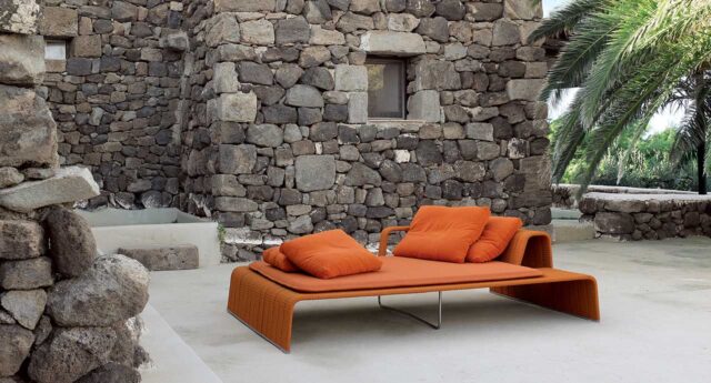 Orangefarbene Outdoor-Lounge mit Kissen vor einem rustikalen Steingebäude und einer Palme, die einen lebendigen, vom luxemburgischen Gartenbau in Deutschland inspirierten Charme versprühen.