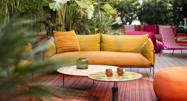 Gelbes und orangefarbenes Burelbach-Sofa im Freien mit bunten Tischen und Stühlen auf einem gestreiften Teppich, umgeben von üppigem Grün.