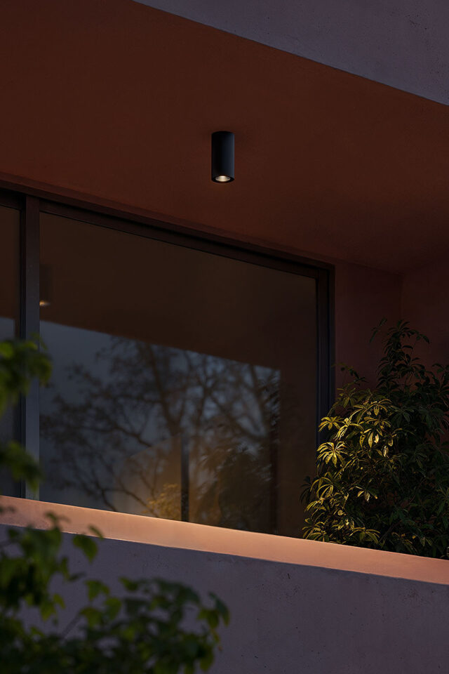 Ein moderner Balkon in der Abenddämmerung von Burelbach mit einer schwarzen Deckenleuchte, einem Glasgeländer, Grünpflanzen und Baumspiegelungen im großen Fenster. Die ruhige, minimalistische Szene unterstreicht den fachkundigen Garten- und Landschaftsbau in Luxemburg und Deutschland.