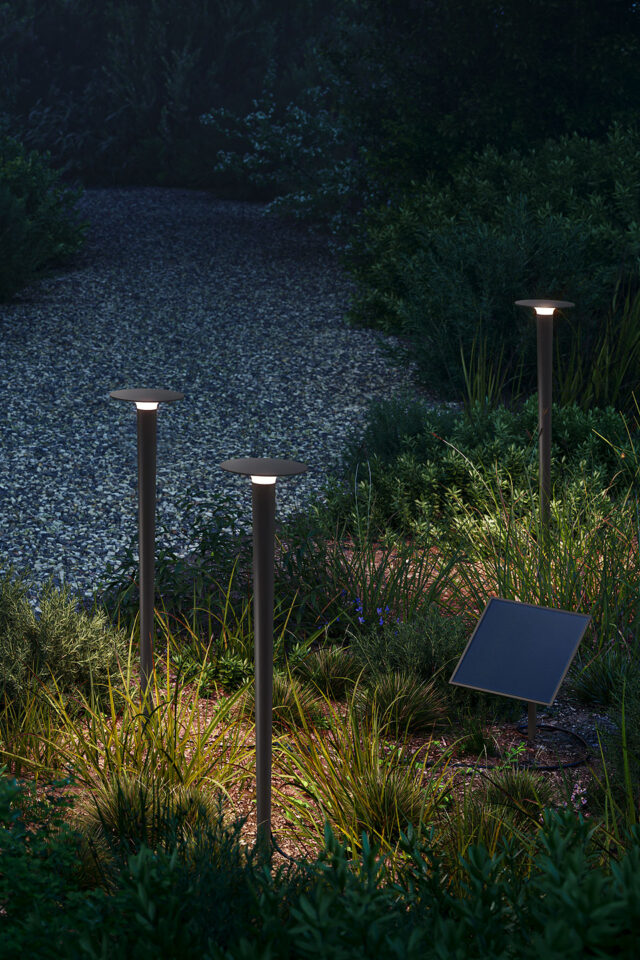 Drei solarbetriebene Gartenleuchten beleuchten nachts einen Kiesweg, der von Gras und Pflanzen umgeben ist. Ein Solarmodul, das in den Garten- und Landschaftsbau integriert ist, fügt sich ganz natürlich in die Szene ein. Burelbach beliefert Luxemburg und Deutschland.