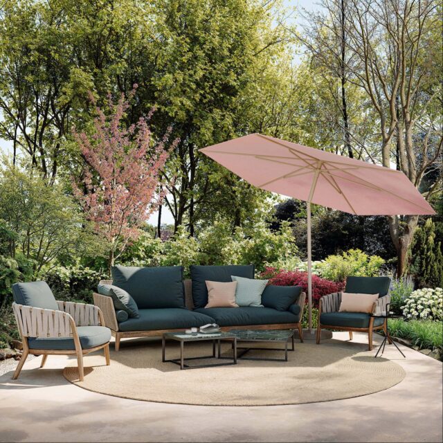 Sitzgruppe im Freien mit dunkelgrünem Sofa, passenden Stühlen, rundem Teppich und Couchtischen unter einem großen rosafarbenen Sonnenschirm - umgeben von üppigem Grün und blühenden Bäumen. Perfekt für Gartendesign-Inspirationen in Deutschland und Luxemburg.