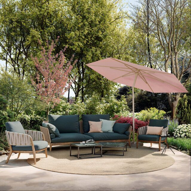 Terrassenset mit Burelbach Gartenmöbeln: ein grünes Polstersofa, zwei Stühle, Couchtische und ein rosafarbener Sonnenschirm auf einem runden Teppich - umgeben von üppigen Bäumen, blühenden Blumen und leuchtenden Gartenpflanzen.