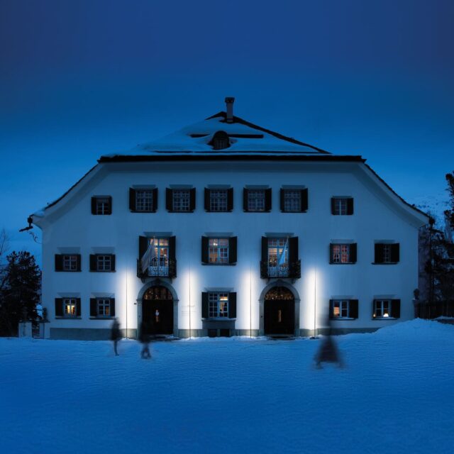 Ein großes, weißes Haus mit grünen Fensterläden wird bei Nacht beleuchtet. Schnee bedeckt den Boden und das Dach, während verschwommene Gestalten davor spazieren gehen - eine einladende Winterszene, die an die Burelbacher Handwerkskunst in Luxemburg und Deutschland erinnert.