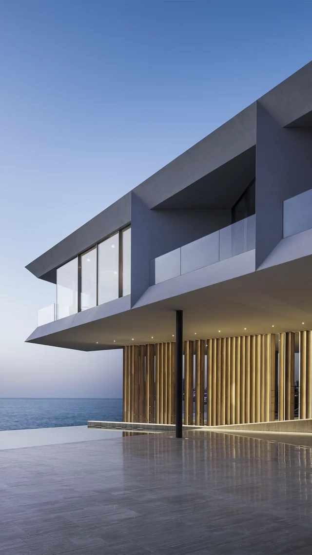 Modernes Strandhaus mit großen Glasfenstern und vertikalen Holzlatten, mit einem freitragenden Obergeschoss, das den Blick auf das Meer bei Sonnenuntergang freigibt, und einer großzügigen, polierten Terrasse, die mit eleganten Gartenmöbeln geschmückt ist, die von Gartentrends aus Luxemburg und Deutschland inspiriert sind.