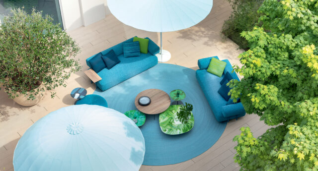 Luftaufnahme einer Terrasse mit blauen Sofas, runden Tischen, blauem Teppich und großen Sonnenschirmen inmitten von Grünpflanzen, die den eleganten Gardendesign-Stil widerspiegeln.