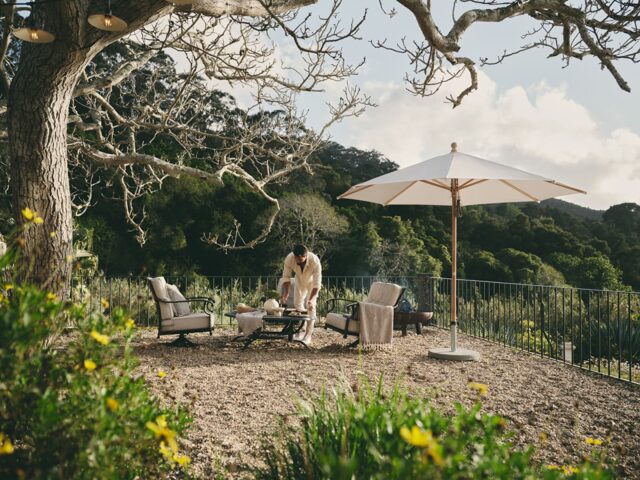 Eine Person steht an einer Feuerstelle in einer gemütlichen Sitzecke im Freien mit Sesseln, einem großen weißen Sonnenschirm und einer Decke - ein Beispiel für Garten- und Landschaftsbau, inspiriert von Gardendesign, umgeben von Bäumen und Grünpflanzen unter einem strahlenden Himmel.