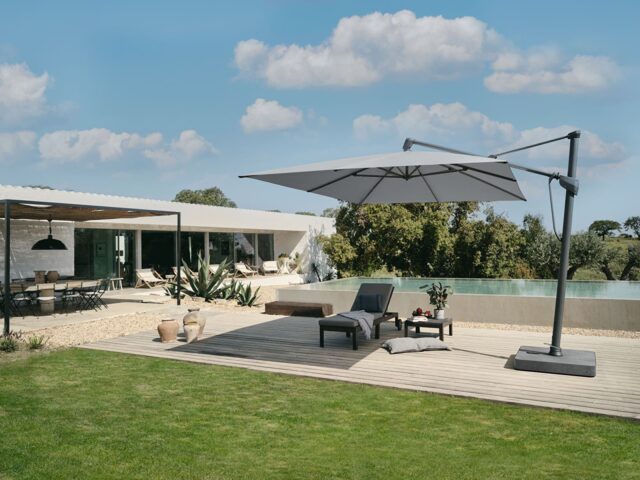 In einem modernen Garten stehen Gartenmöbel auf einer Holzterrasse mit zwei Loungesesseln, einem Beistelltisch und einem großen freitragenden Sonnenschirm in der Nähe eines Infinity-Pools. Elegantes Handwerk erstrahlt in dieser modernen Oase unter dem blauen Himmel.