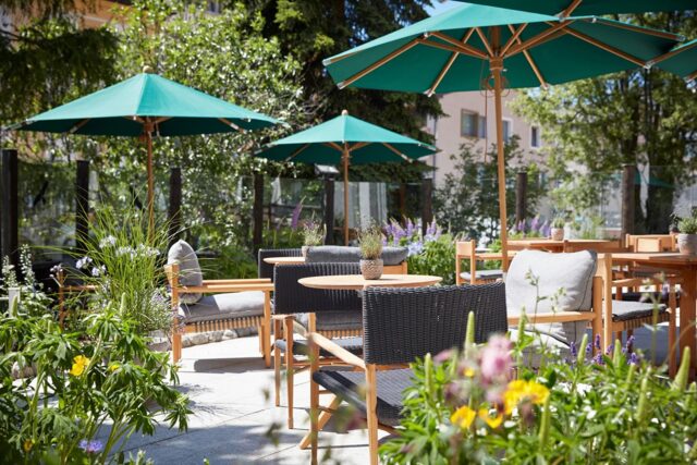 Außenterrasse mit Burelbach Gartenmöbeln mit Holztischen und -stühlen, grauen Kissen, grünen Sonnenschirmen und Topfpflanzen. An einem sonnigen Tag umgeben bunte Blumen und Grünpflanzen den Sitzbereich.