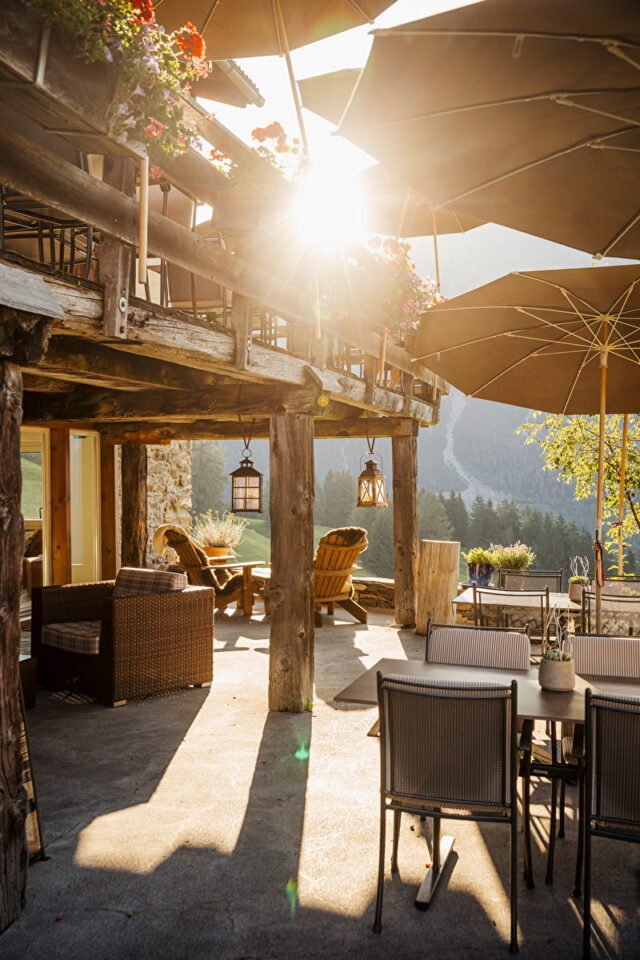 Eine sonnenbeschienene Außenterrasse in einer rustikalen Holzhütte in Deutschland und Luxemburg mit handwerklich inspirierten Tischen, Sitzgelegenheiten aus Korbgeflecht, Topfblumen und großen Sonnenschirmen - mit Blick auf eine malerische Berglandschaft mit Bäumen dahinter.