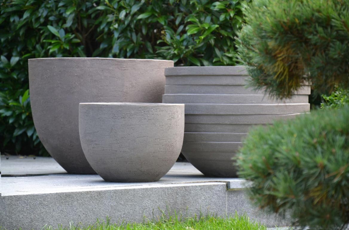Drei große, graue, strukturierte Pflanzgefäße in verschiedenen Formen und Größen stehen auf einer Steinterrasse und zeigen Burelbachs Kompetenz in Gartendesign und Garten- und Landschaftsbau inmitten von üppigen grünen Sträuchern und Gras.