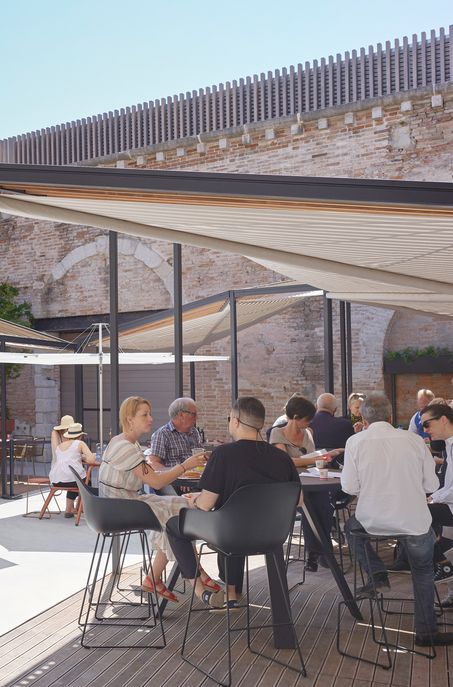 Die Menschen sitzen an Tischen im Freien unter modernen Schattenspendern und genießen das Essen und die Gespräche an einem sonnigen Tag. Gartenmöbel und Steinmauern schaffen eine entspannte, ungezwungene Atmosphäre, die perfekt ist, um sich mit Freunden zu treffen.