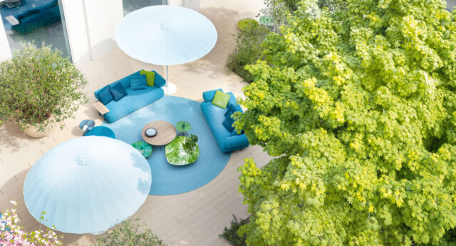 Eine Außenterrasse mit blauen Sofas, Sonnenschirmen und einem runden Teppich neben einem großen grünen Baum, von oben gesehen - ein stilvolles Gardendesign von Burelbach.