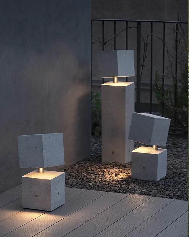 Drei moderne Außenlampen aus Beton mit würfelförmigen Sockeln und außermittig angeordneten quadratischen Aufsätzen geben warmes Licht ab. Zwei der Lampen sind ideal für Gardendesign und stehen auf einer Holzterrasse, während die größte Lampe nachts auf Kies in der Nähe eines Metallgeländers und spärlicher Pflanzen steht.