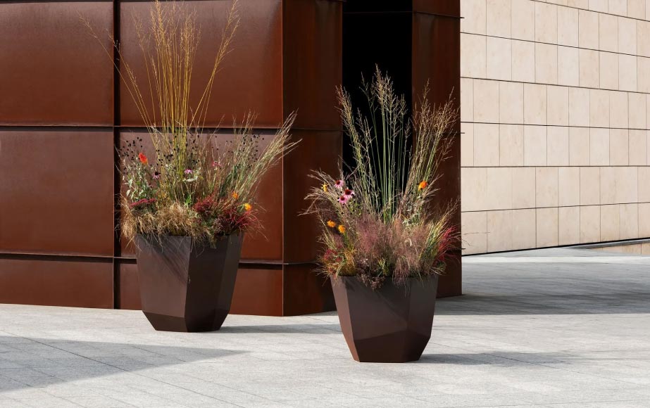Zwei große, moderne geometrische Pflanzgefäße, die mit Burelbach Handwerk gefertigt und mit hohen Gräsern und Wildblumen gefüllt wurden, stehen auf einer gepflasterten Fläche neben modernen Gebäuden und werten den Raum wie elegante Gartenmöbel auf.