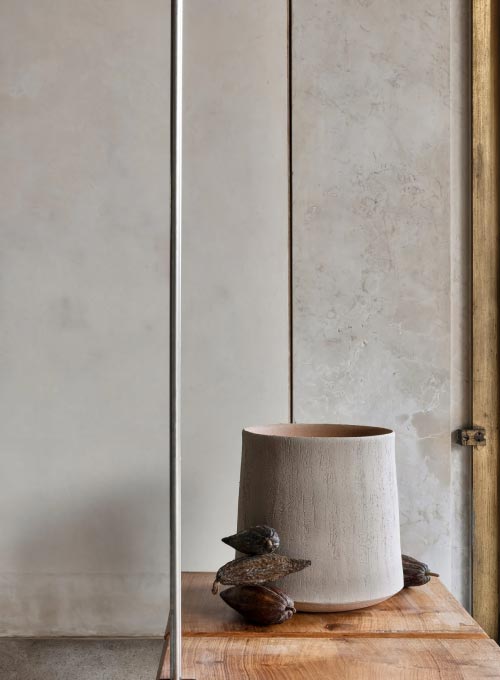 Eine minimalistische Szene mit einer strukturierten Keramikvase und zwei kleinen hölzernen Vogelskulpturen, die feines Handwerk widerspiegeln, angeordnet auf einer hölzernen Oberfläche vor einer hellen, strukturierten Wand.