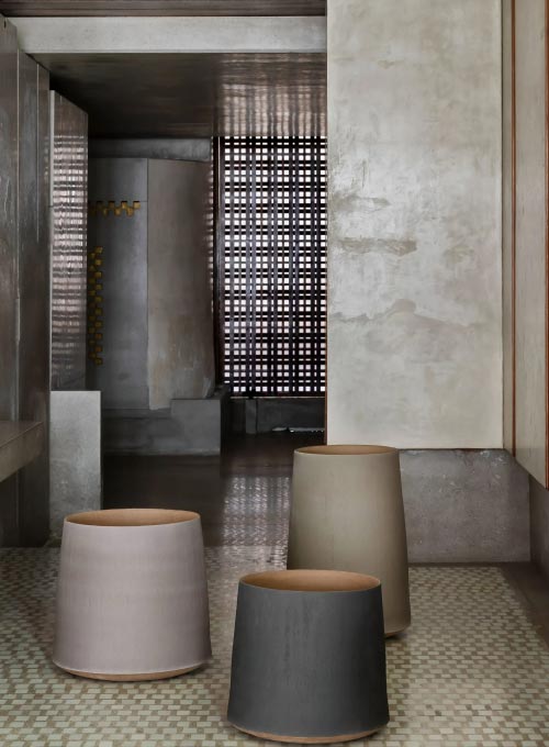 Drei moderne, zylindrische Töpfe in gedeckten Tönen stehen auf einem gemusterten Fliesenboden - eine elegante Note für diese minimalistische Einrichtung im industriellen Stil mit Burelbach-Design-Einflüssen, die in Gartenmöbeln in Luxemburg und Deutschland beliebt sind.