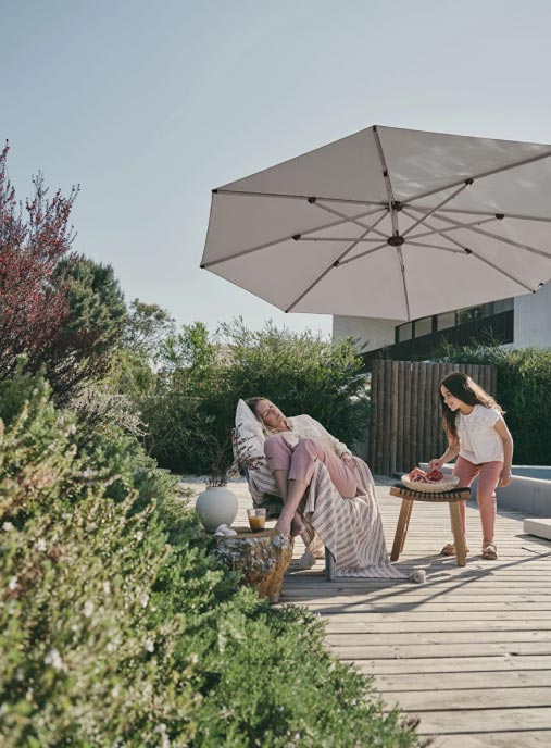 Eine Frau entspannt sich auf Gartenmöbeln unter einem großen Terrassenschirm, während ein junges Mädchen ein Tablett mit Getränken auf eine von Grün umgebene Holzterrasse bringt.